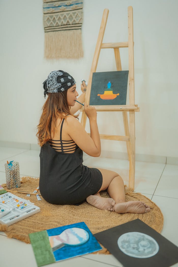Young woman in headscarf painting on easel at home, showcasing creativity and artistic skill.