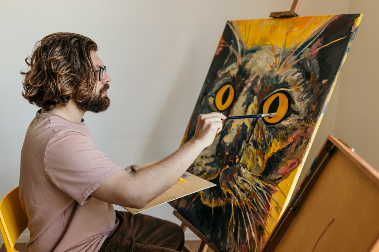 Man artist painting a colorful cat portrait on canvas indoors.