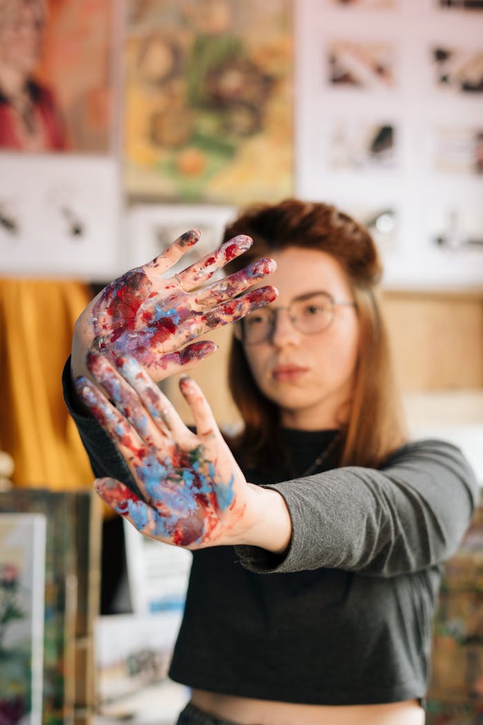 about-us-02 Young female artist showcasing her colorful painted hands in a vibrant art studio setting.